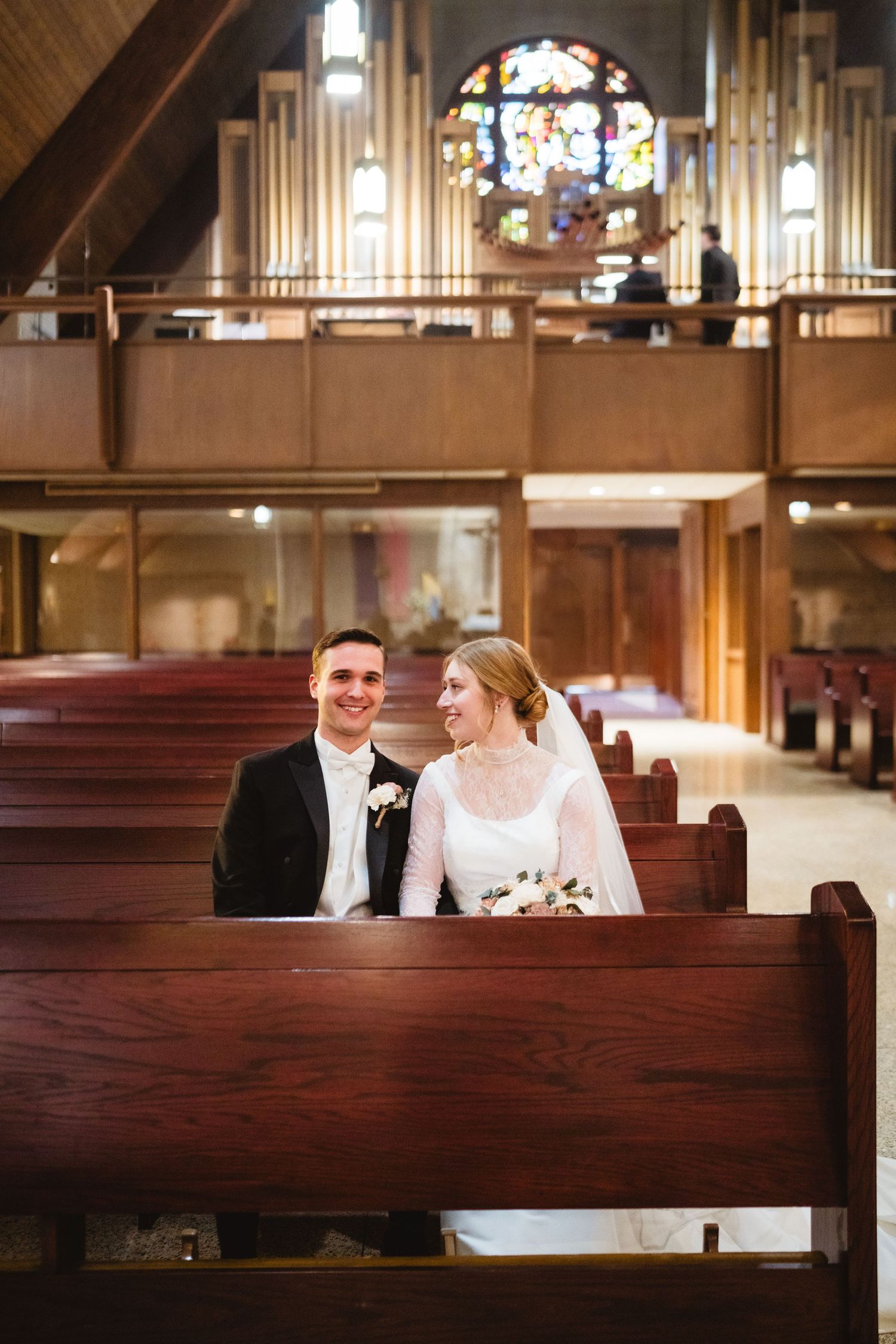 A couple sits together in a wooden church pew with a stained glass window and organ pipes visible in the background.