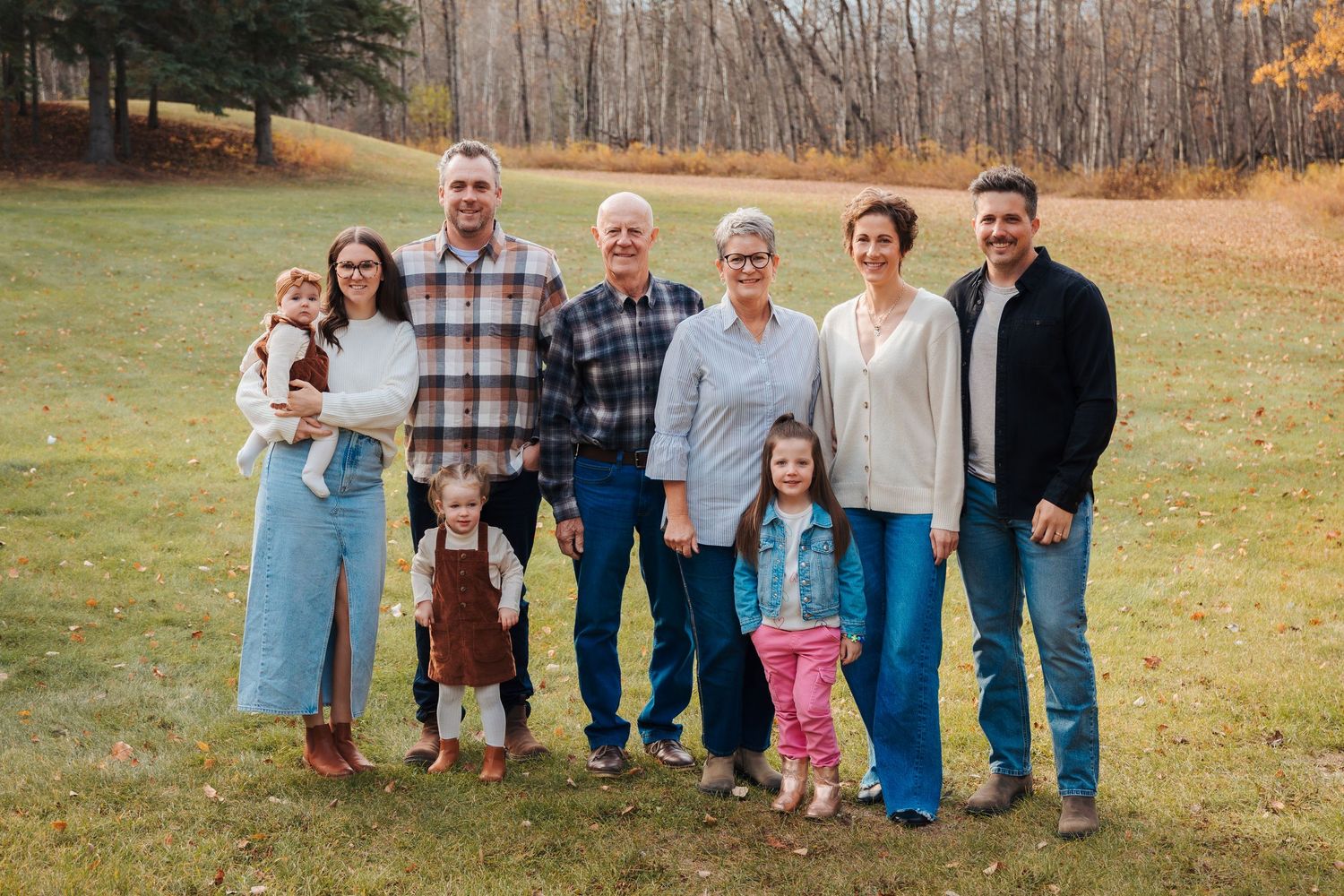 A multigenerational family of eight poses together smiling in a grassy autumn outdoor setting.