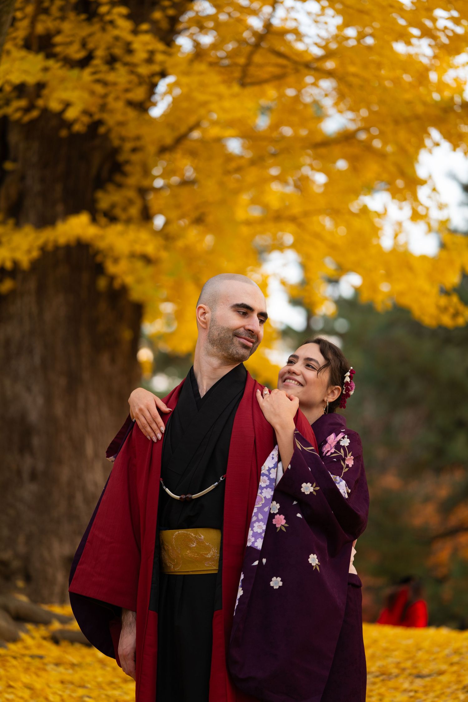 Smiling couple in traditional Japanese kimono embracing under a golden ginkgo tree in autumn.