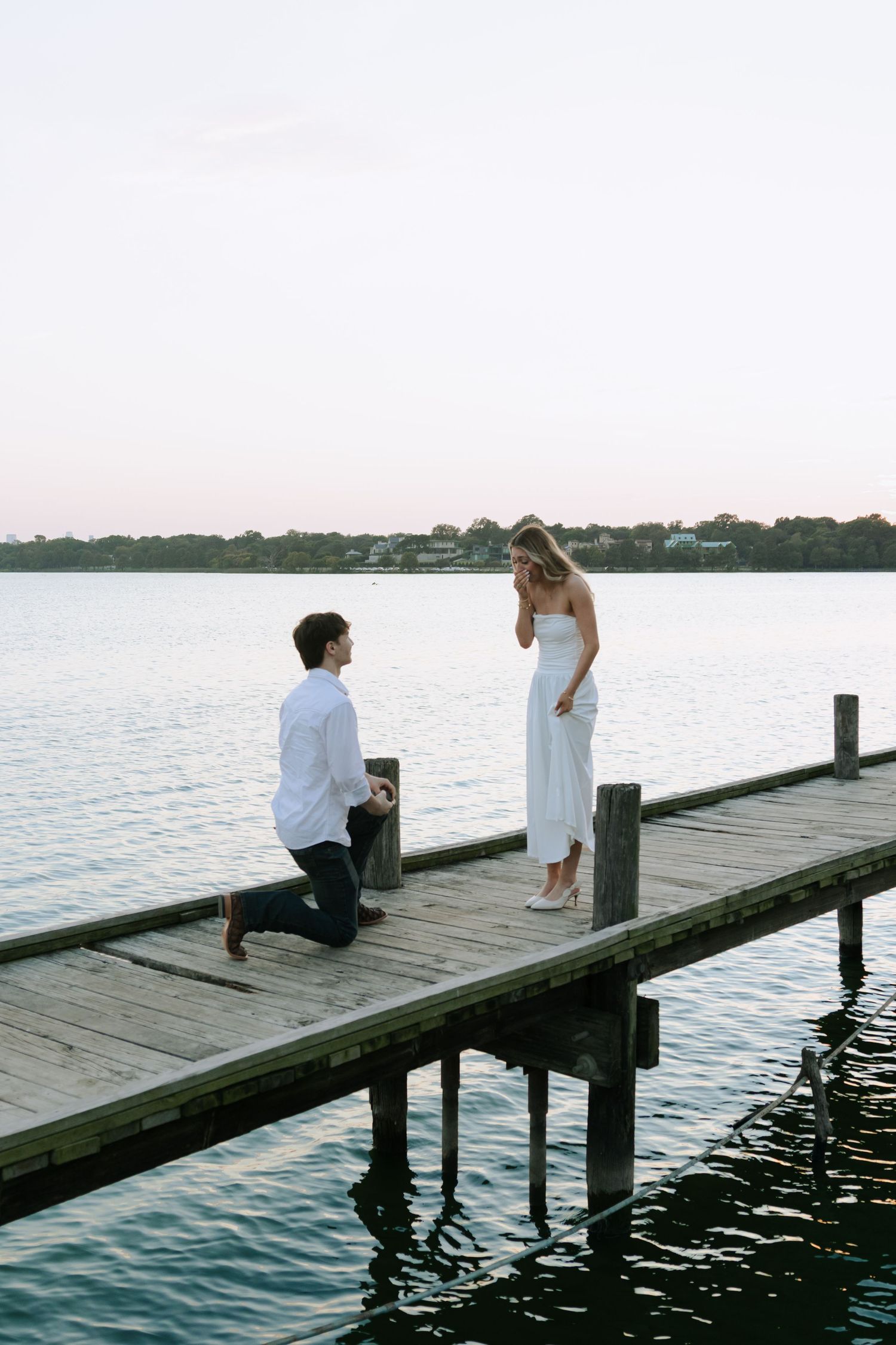 A romantic marriage proposal takes place on a wooden dock over calm waters at sunset.