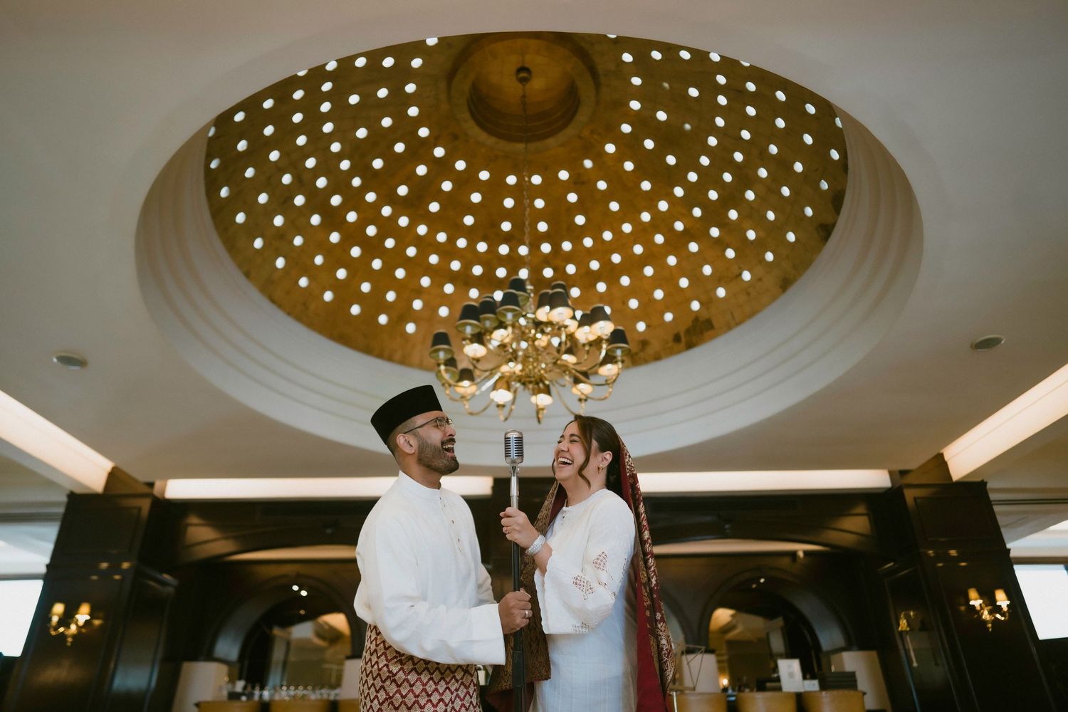Malay couple in traditional attire standing beneath an ornate golden domed ceiling in a luxury hotel.