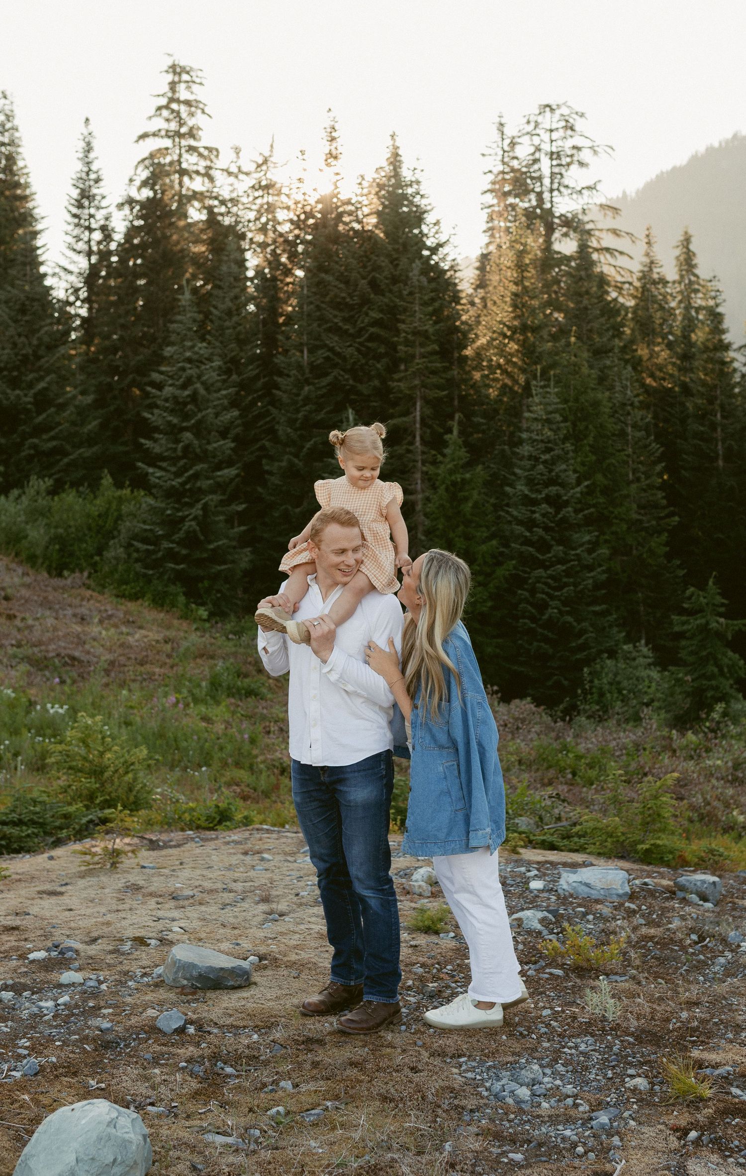 Couple shares tender moments at sunset in a mountain forest setting with evergreen trees and rocky terrain.
