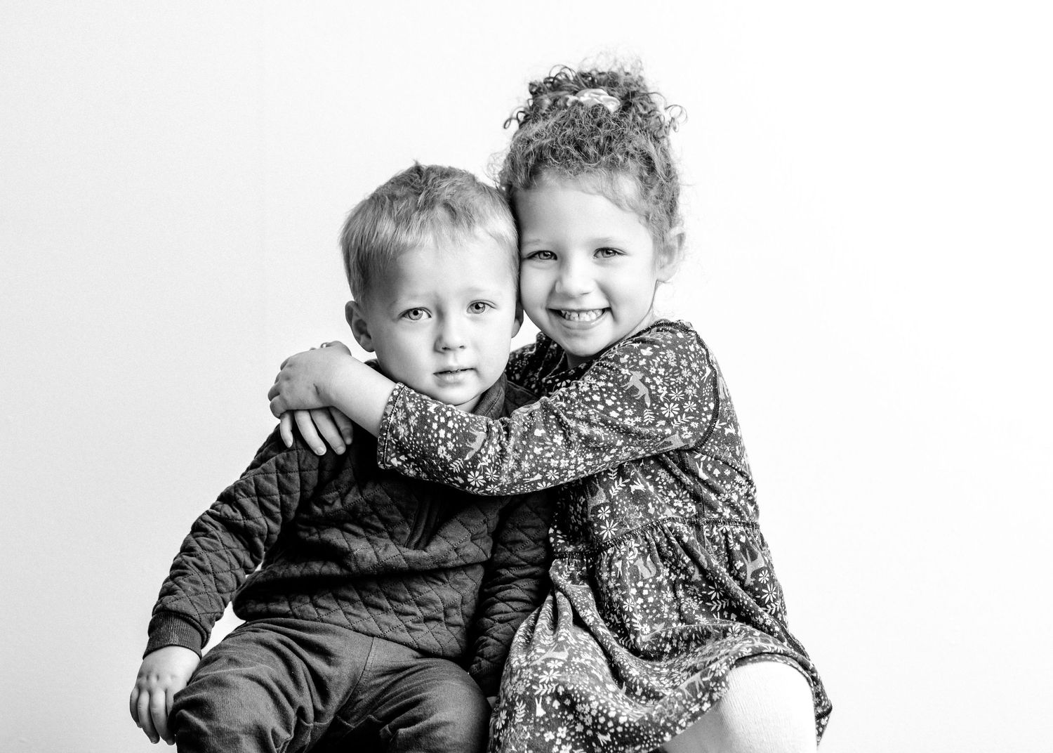 Two young children in winter coats and scarves share a playful embrace outdoors.