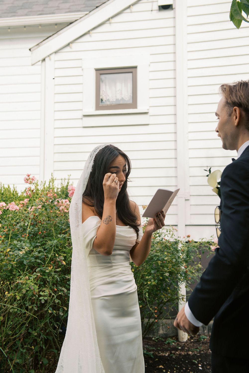 Candid sequence of a bride and groom sharing a playful first look moment in front of a white house with flowering bushes.