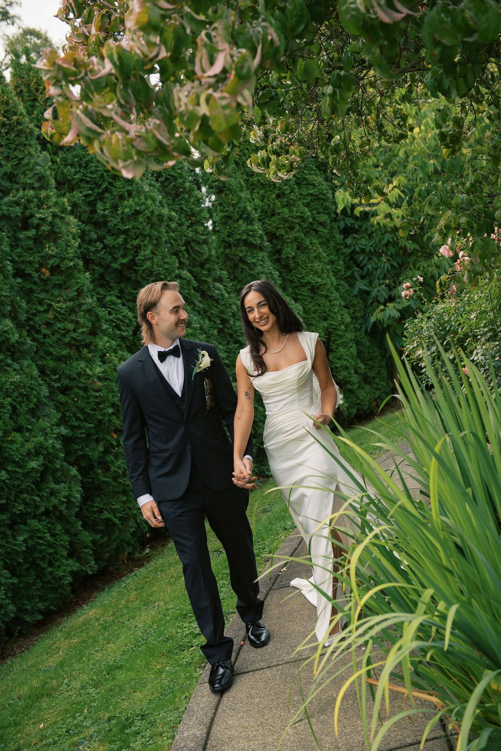 A newlywed couple walks hand in hand through a garden path surrounded by lush greenery.