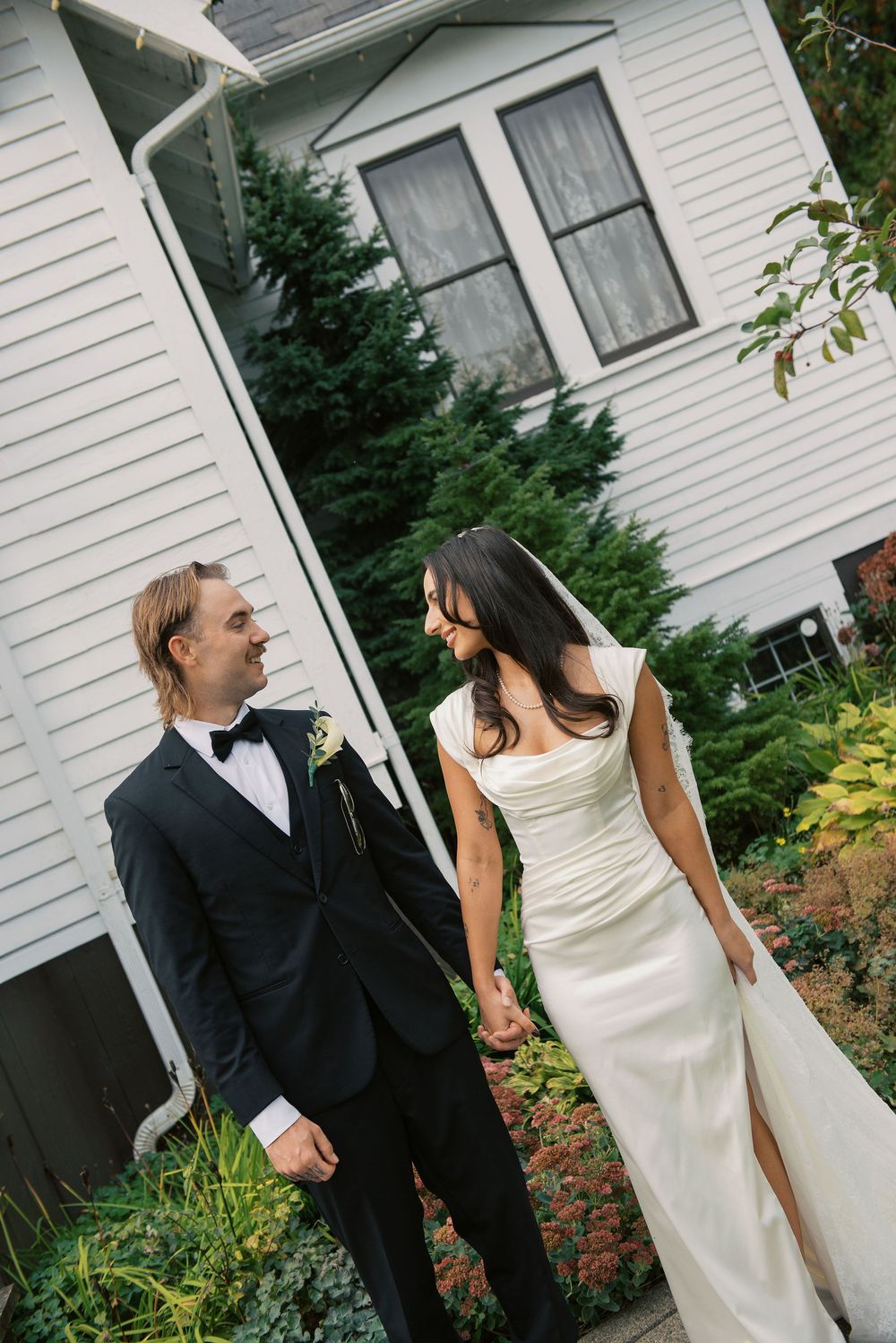 A couple in wedding attire share a romantic moment in front of a white house surrounded by greenery.
