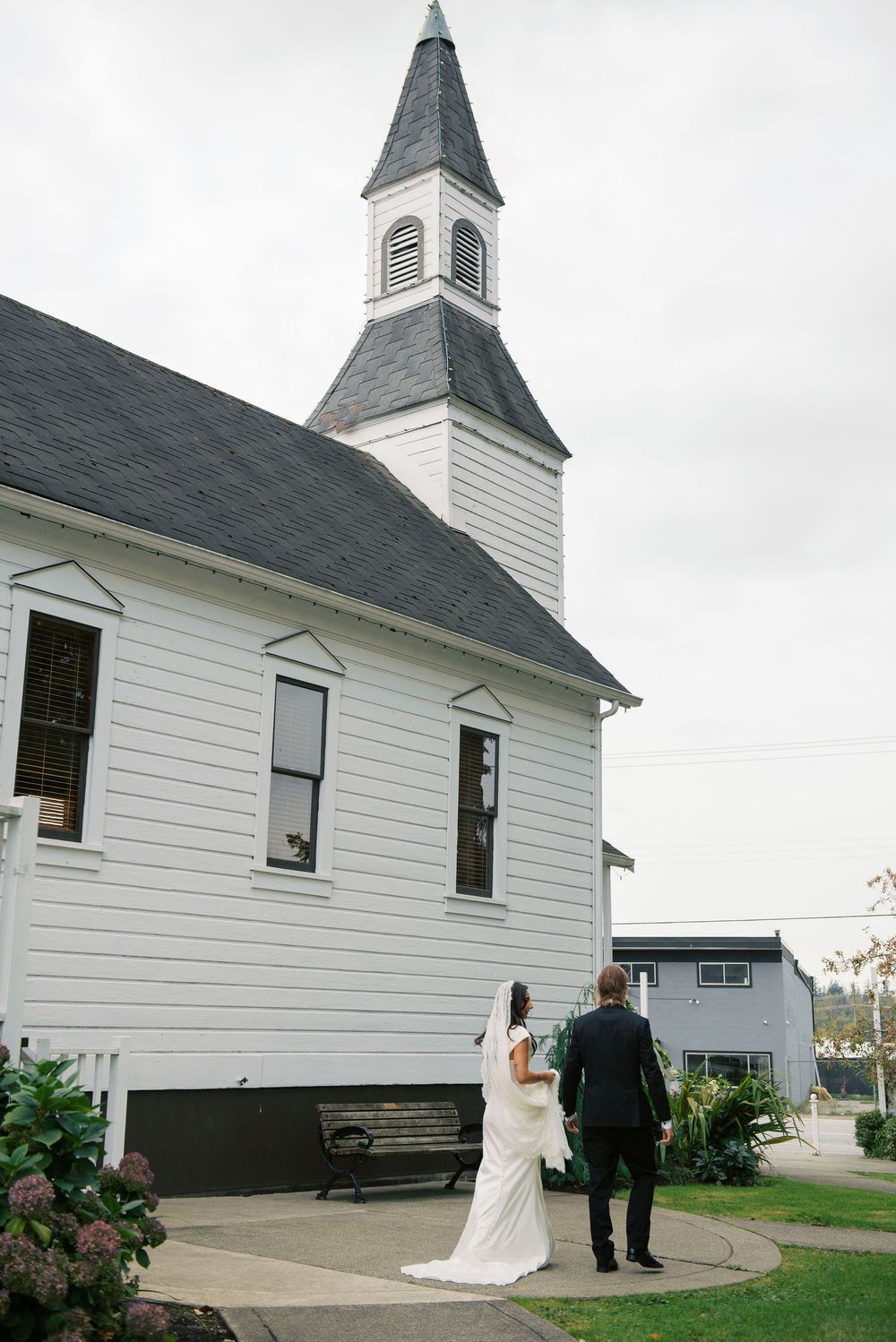A white country church with a tall steeple stands against an overcast sky with a couple on the steps.