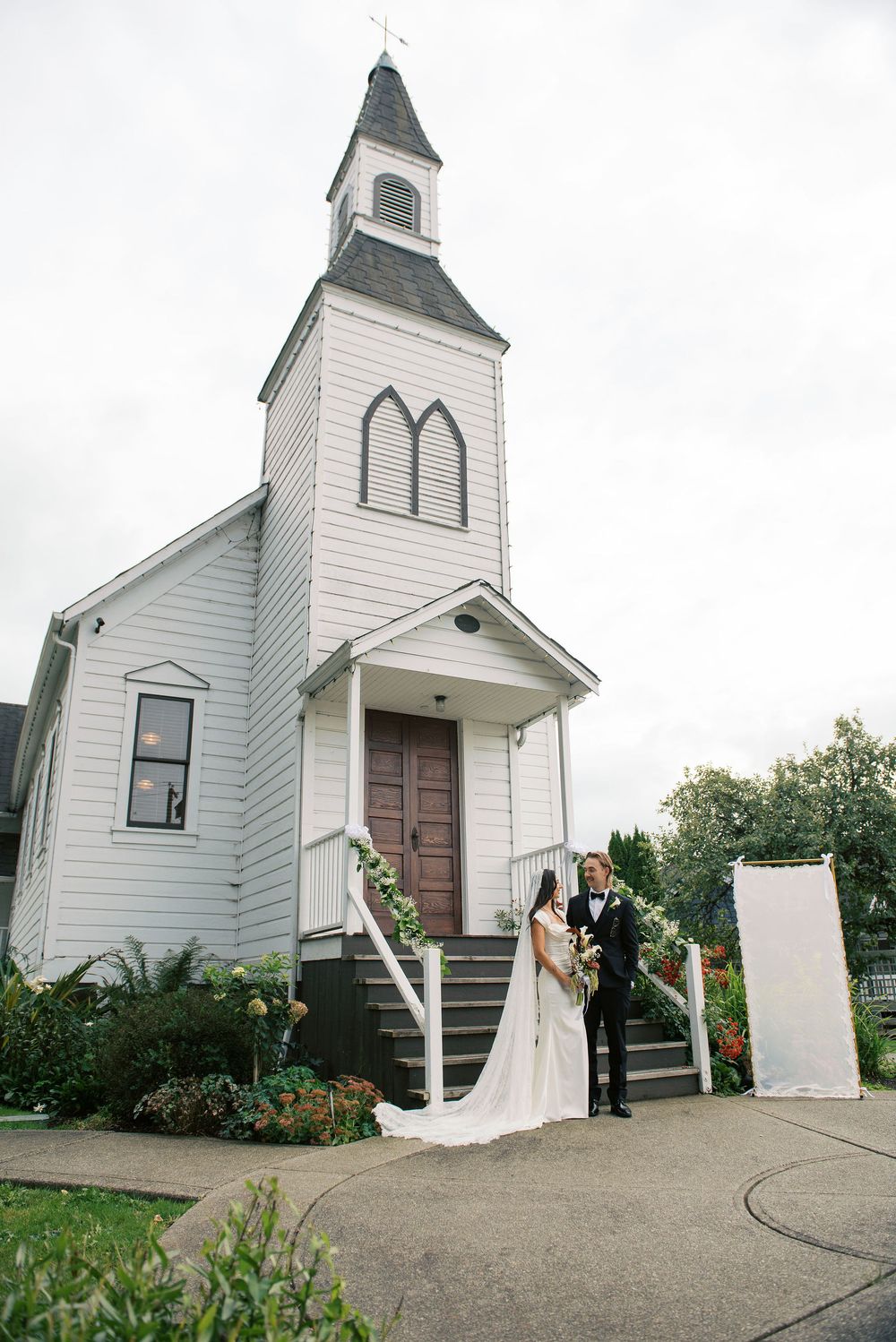 A charming white country church with a tall steeple serves as backdrop for a wedding ceremony.