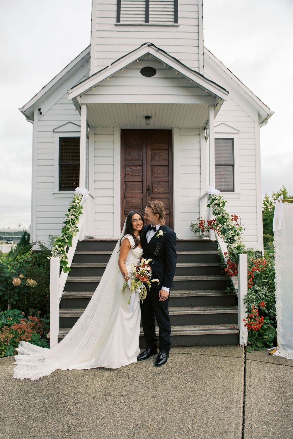 A couple in wedding attire stands on the steps of a charming white chapel with floral decorations.