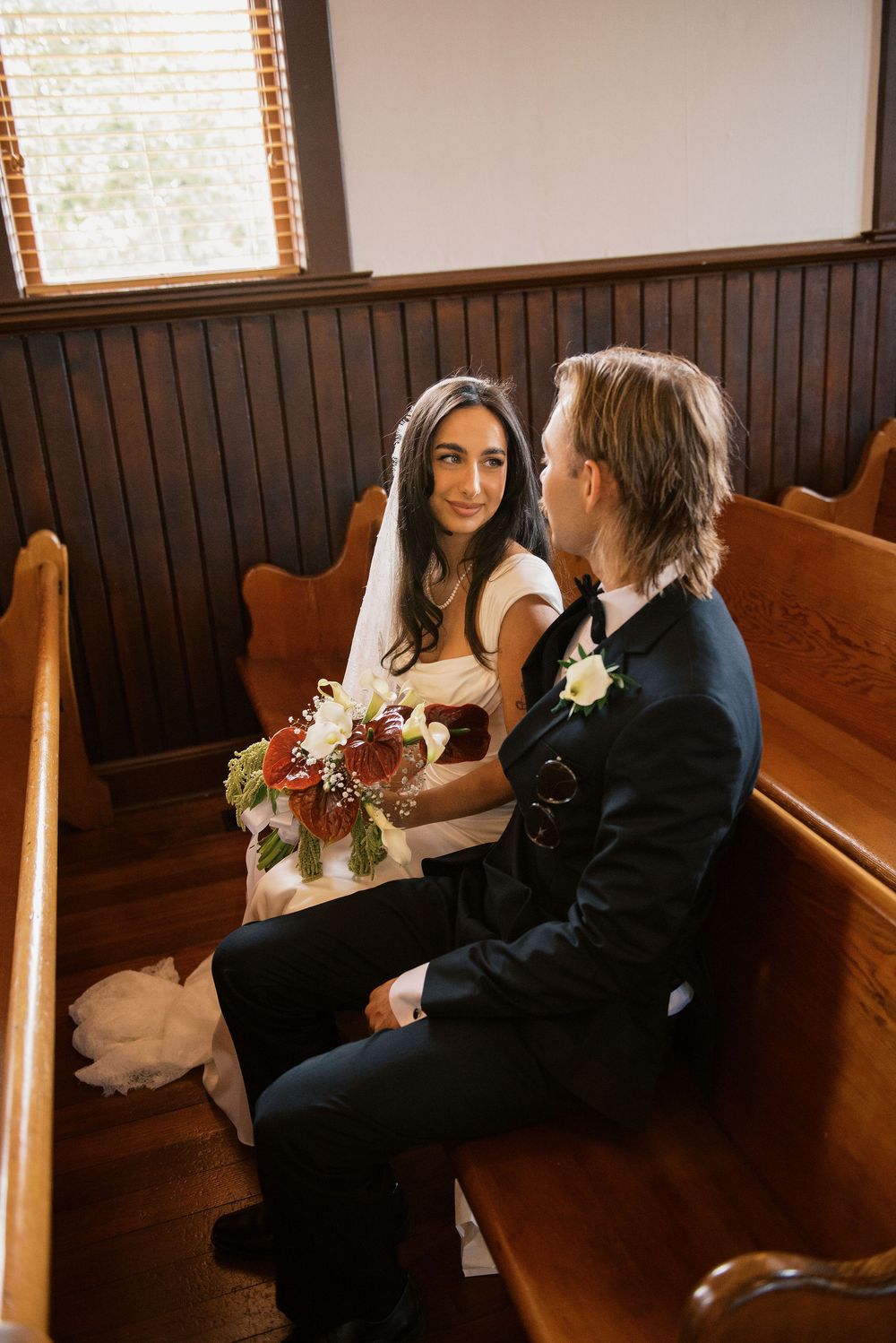 A couple sits together on wooden church pews during their intimate wedding ceremony.