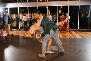 People dance together in a social dance class setting with wooden floors and large windows at night.