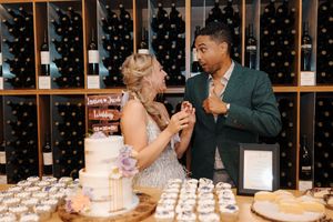 A couple shares a romantic moment while tasting wedding cake samples at an elegant venue with wine bottles in the background.