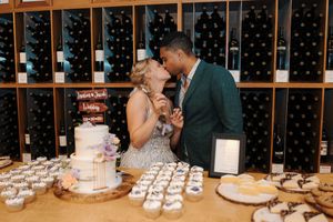 Wedding reception dessert table with cake and cupcakes displayed against a wall of illuminated wine bottles.