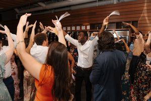 Wedding guests dance and celebrate with raised arms on a crowded dance floor at an indoor reception venue.