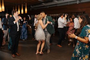 Large group of wedding guests dancing and mingling in a wood-paneled reception venue with pendant lighting.