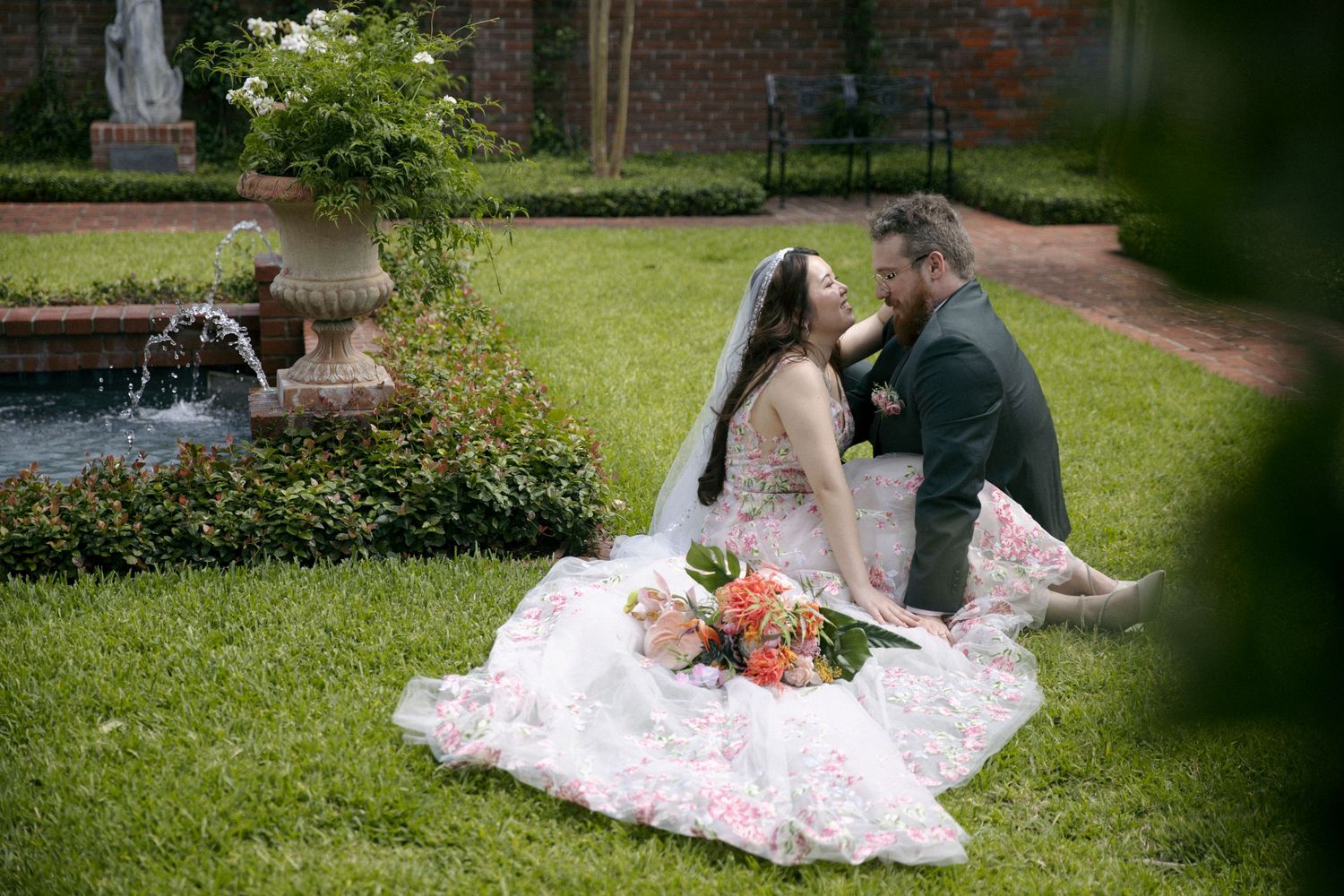 A couple sits on a pink blanket in a garden setting with a fountain and brick building in the background.
