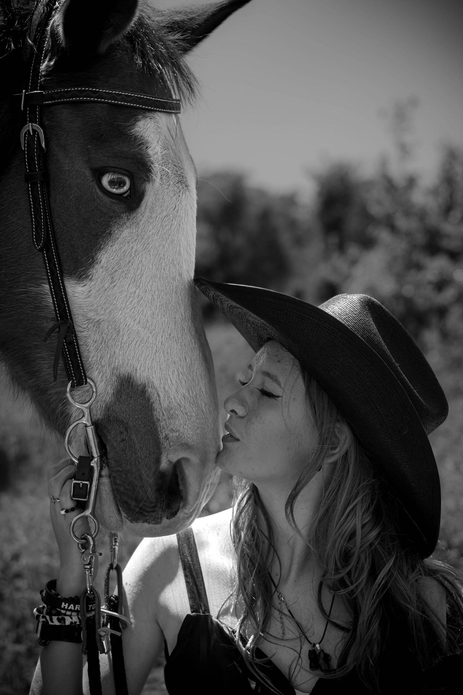 A black and white photograph shows a tender moment between a woman in a cowboy hat and her horse in natural lighting.