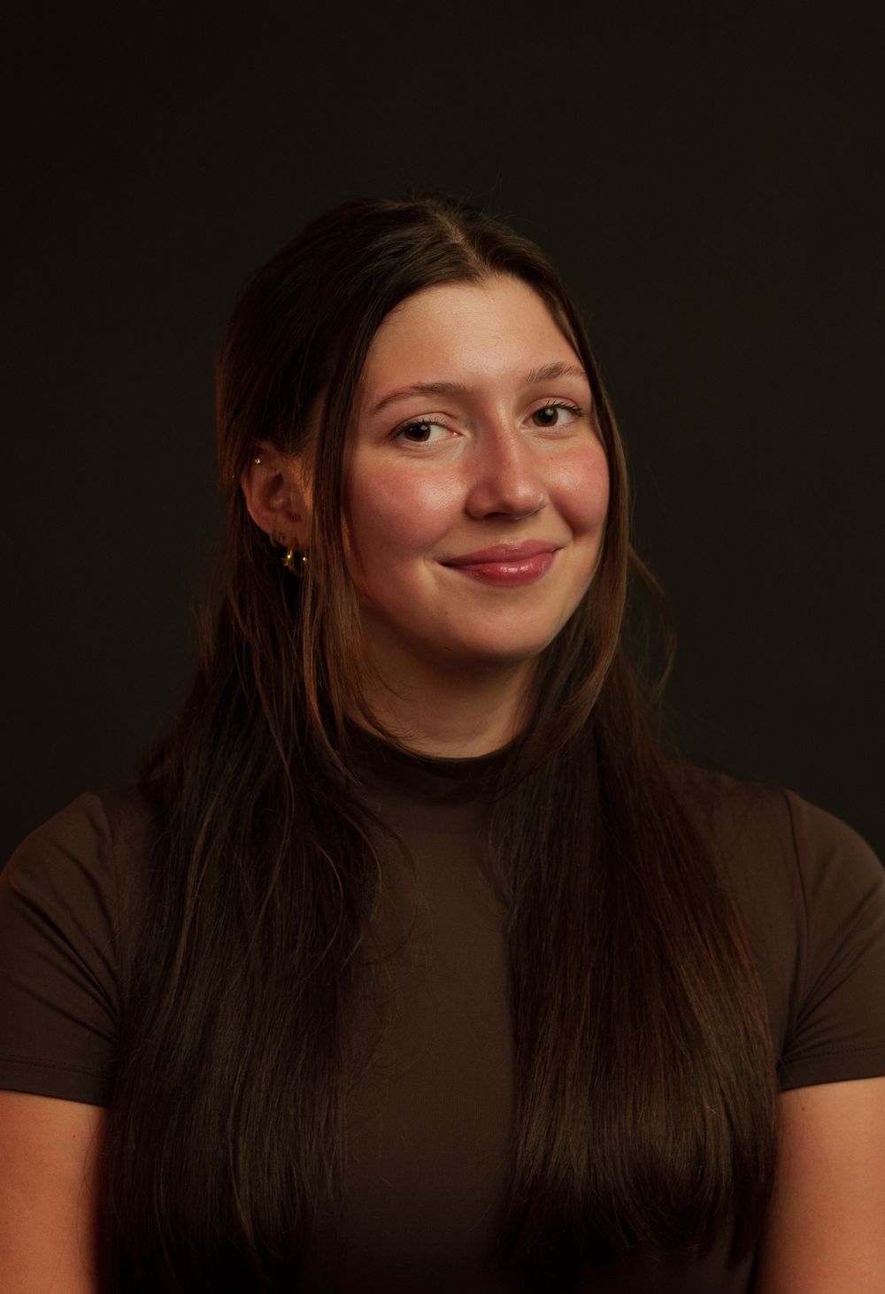 Portrait of someone wearing a brown turtleneck against a dark background with long dark hair and a warm smile.