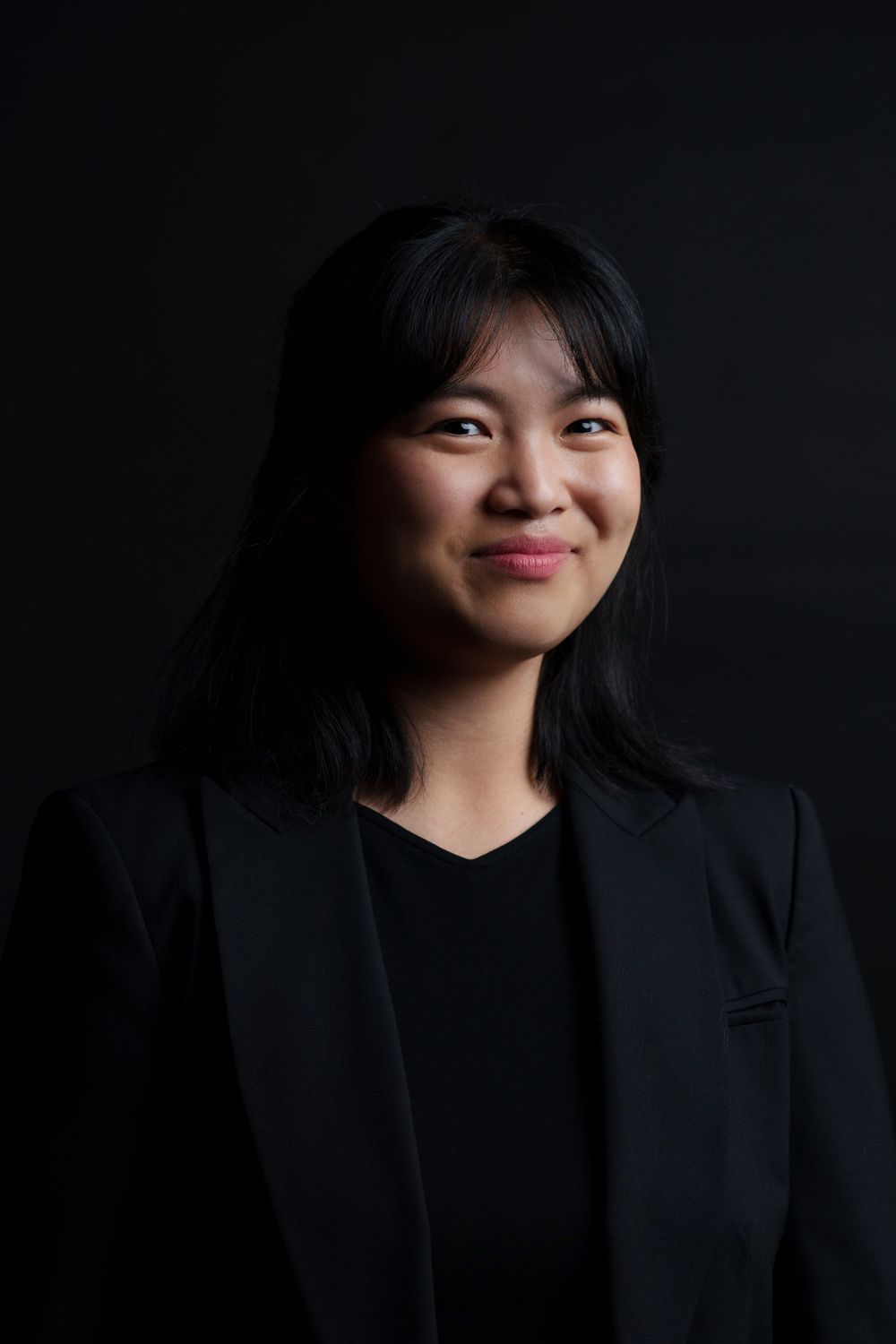 Professional headshot portrait of someone in a black blazer smiling warmly against a dark background.