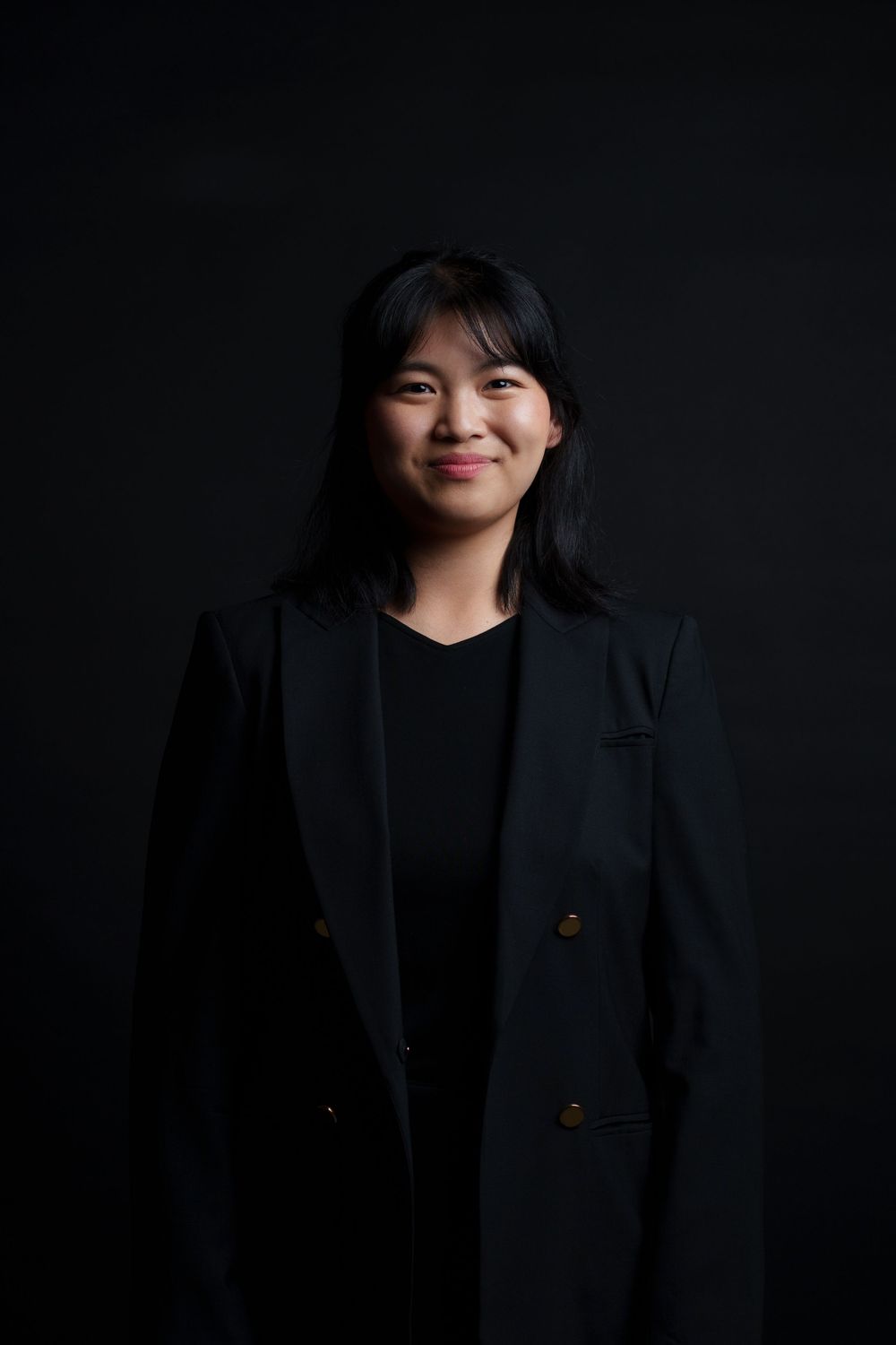 Professional studio portrait against dark background shows person in black blazer with warm expression.