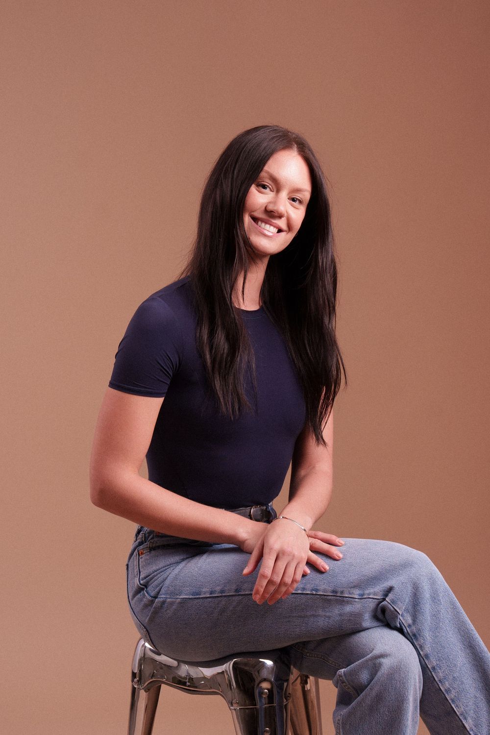 A person in a navy t-shirt and jeans sits casually on a wooden stool against a brown backdrop.