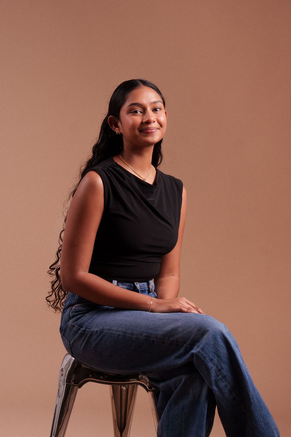 Someone in a black sleeveless top and blue jeans sits casually on a stool against a brown studio backdrop.
