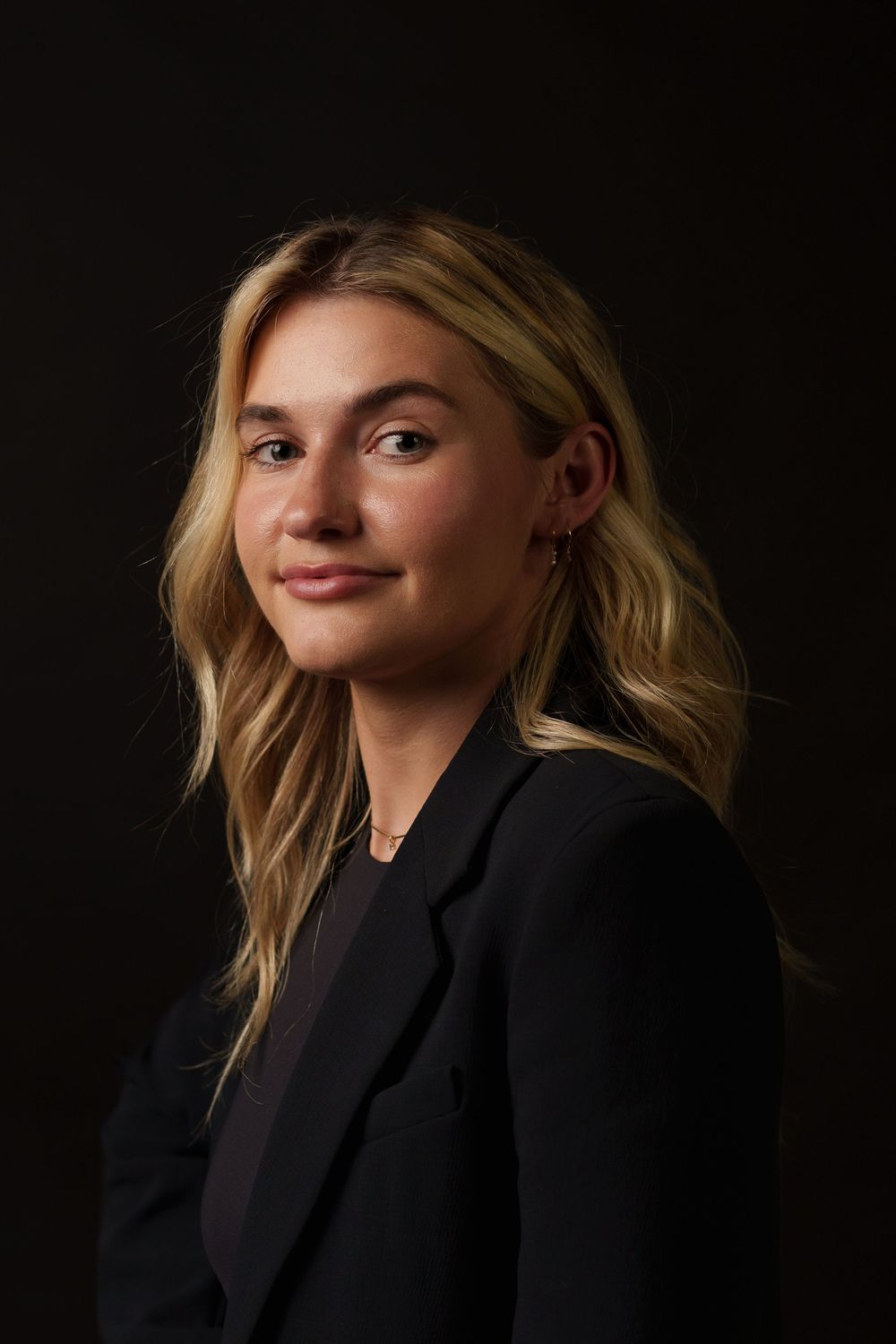 Professional portrait of someone wearing a black blazer against a dark background, with wavy blonde hair.