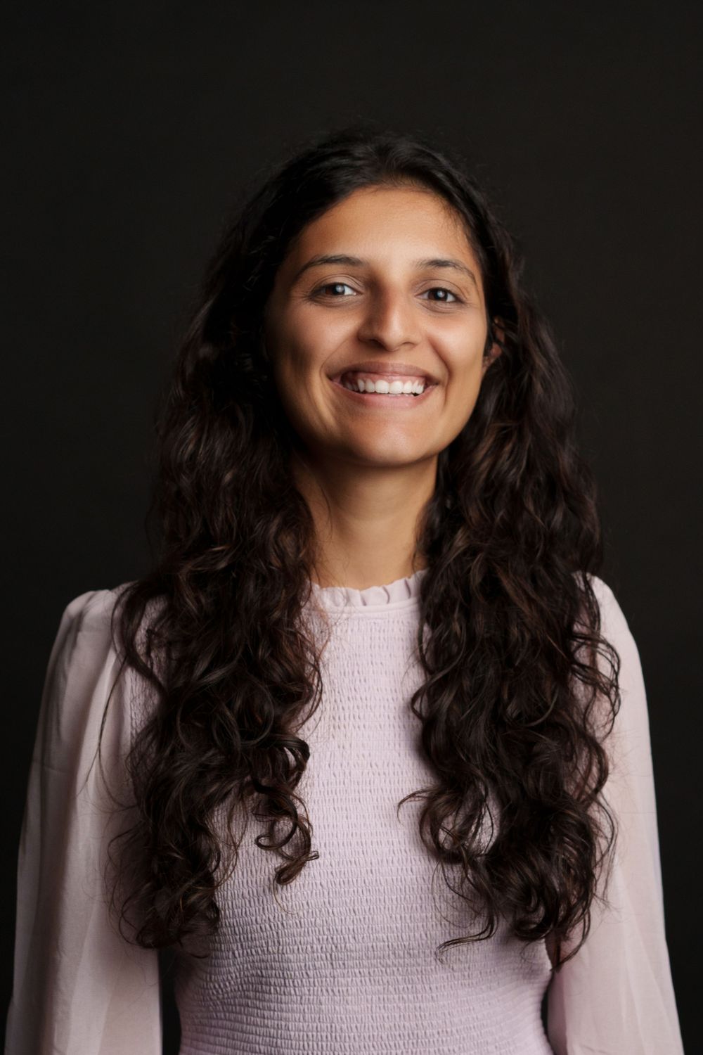 Professional portrait of a person with long dark wavy hair wearing a light pink sweater against a dark background.