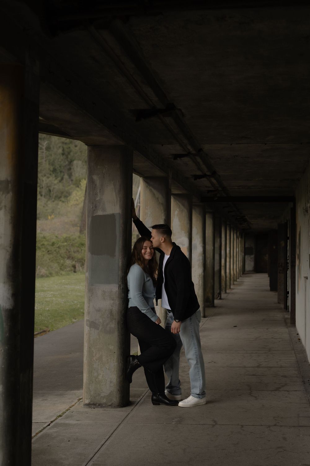 A couple shares an intimate moment in a dramatic archway corridor with moody lighting and concrete pillars.