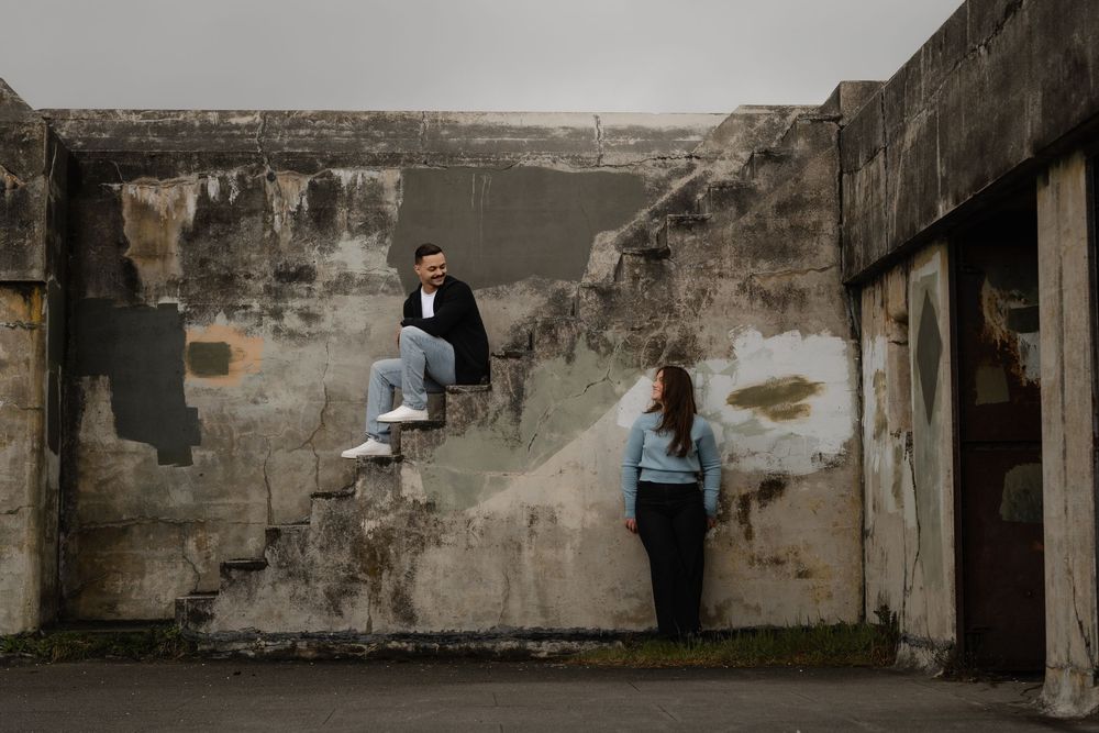 Two people in casual clothing stand against weathered concrete walls in an urban setting with overcast sky.
