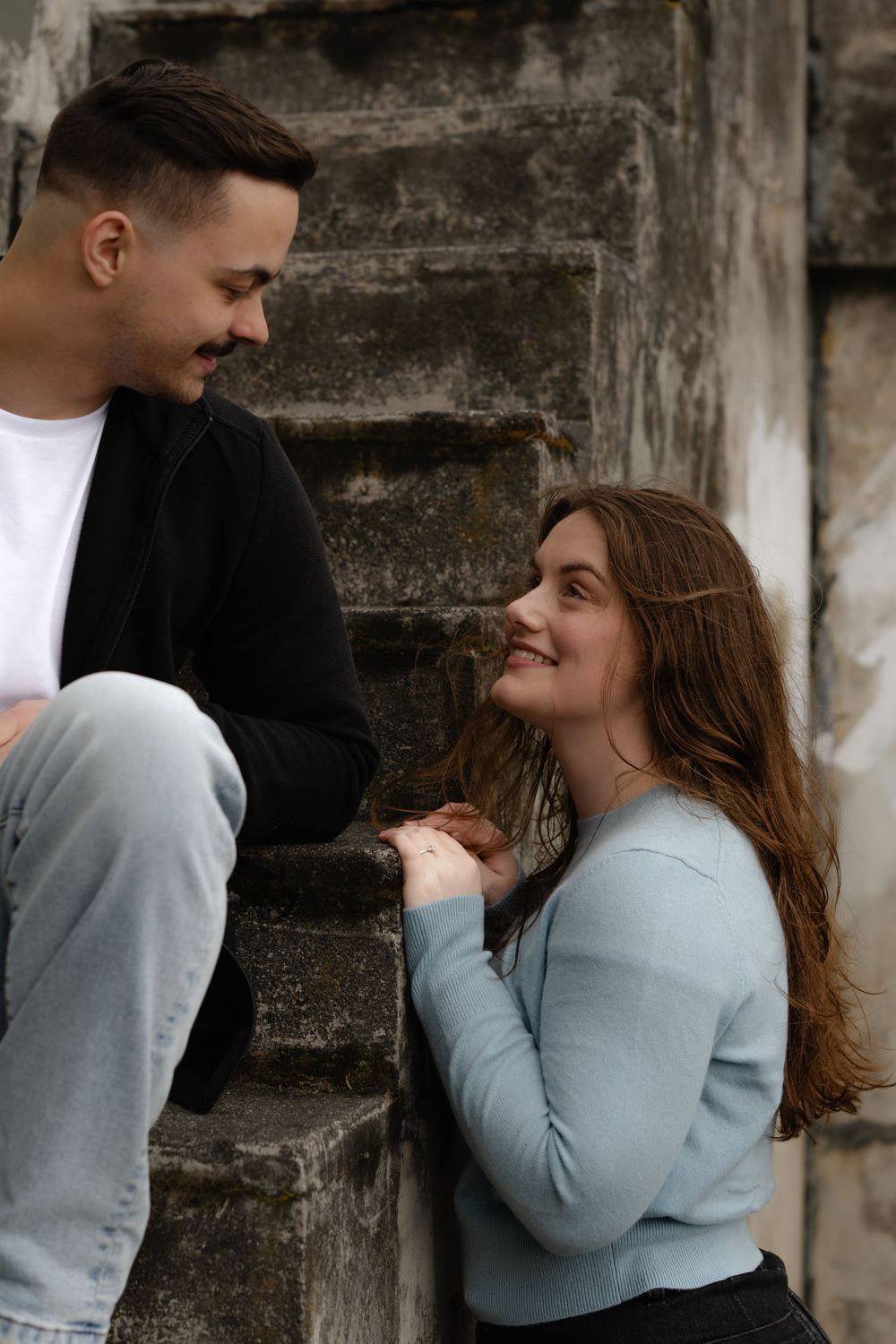 A couple shares a tender moment sitting on concrete steps, with the person in light blue smiling up at their companion.