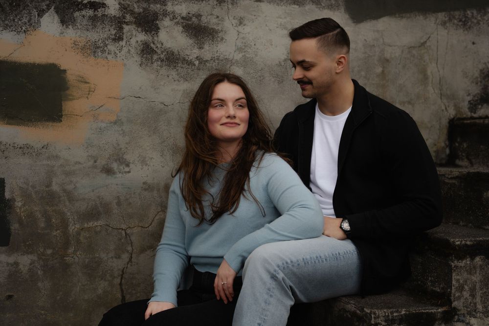 A couple in casual attire sits together against a textured concrete wall during an intimate portrait session.