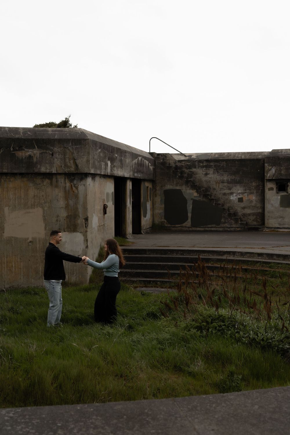 Two people explore old concrete military bunker ruins against an overcast sky.