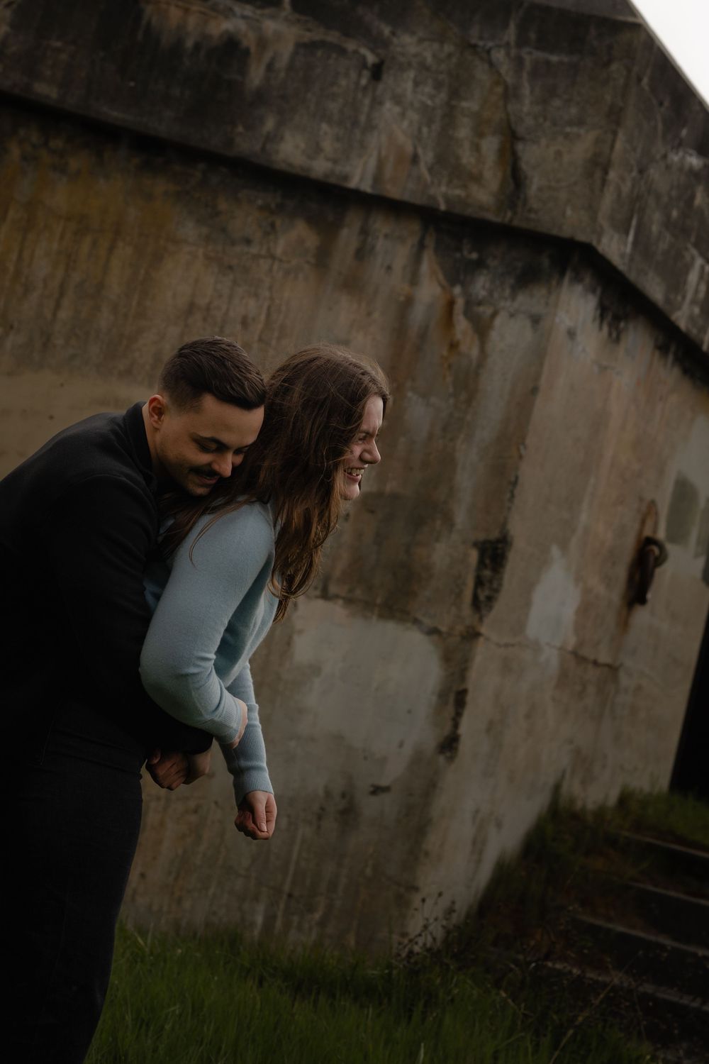 A couple sharing a joyful moment against an industrial concrete wall backdrop in moody lighting.