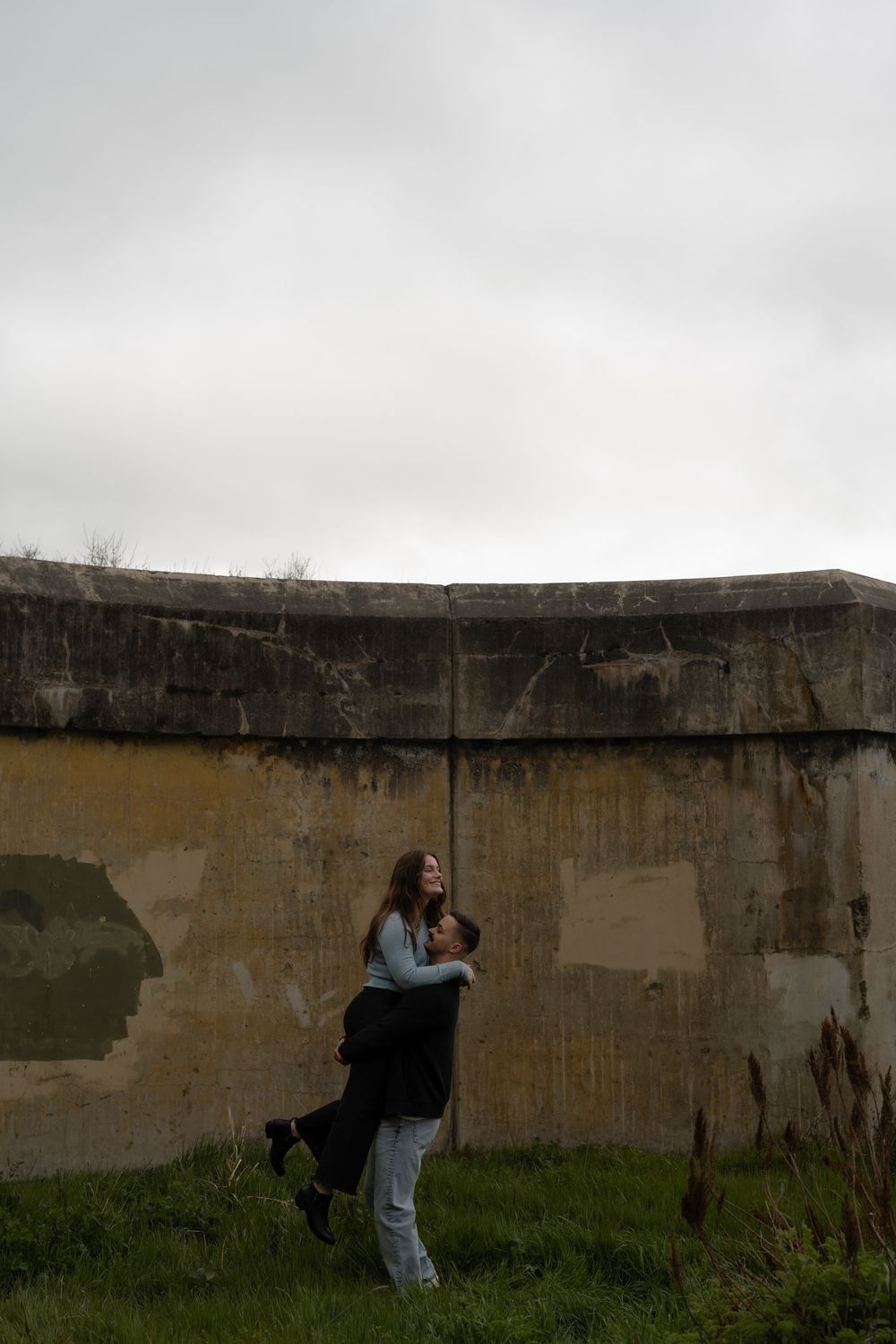 Two figures embrace in front of a weathered concrete wall on a cloudy day with grass in the foreground.