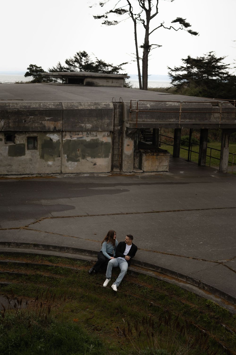 A couple sits together on a grassy slope near an old stone wall at sunset, with trees silhouetted against a misty sky.