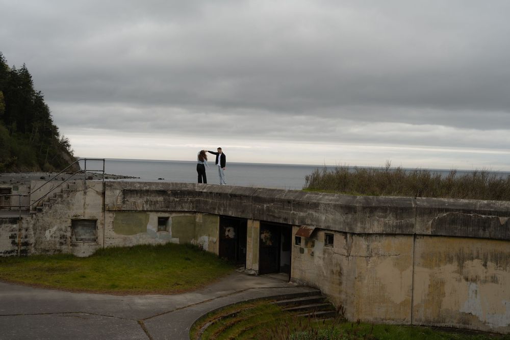 Two silhouetted figures stand on concrete military bunker ruins overlooking a misty ocean coastline under cloudy skies.