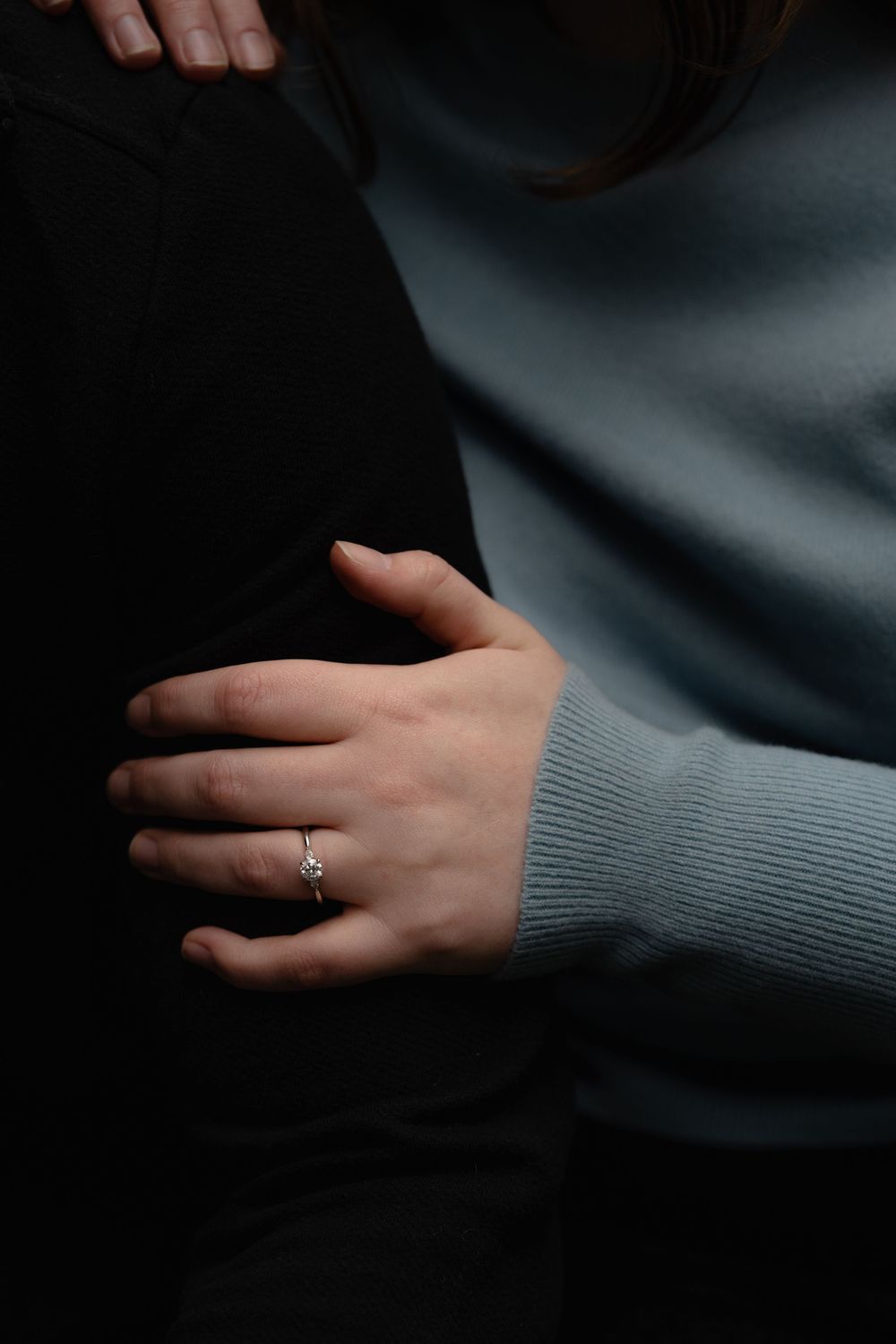 Close-up of a hand wearing a gray sweater sleeve and delicate engagement ring against a dark background.