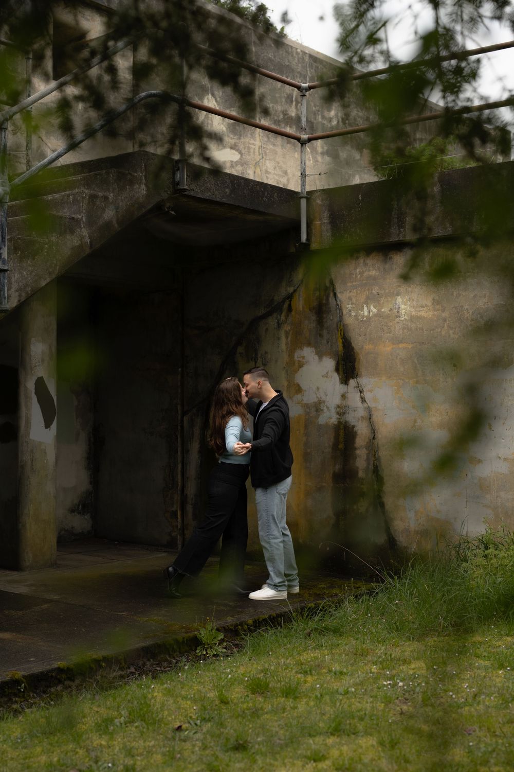 A couple embraces beneath a concrete military bunker structure framed by green foliage on the edges.