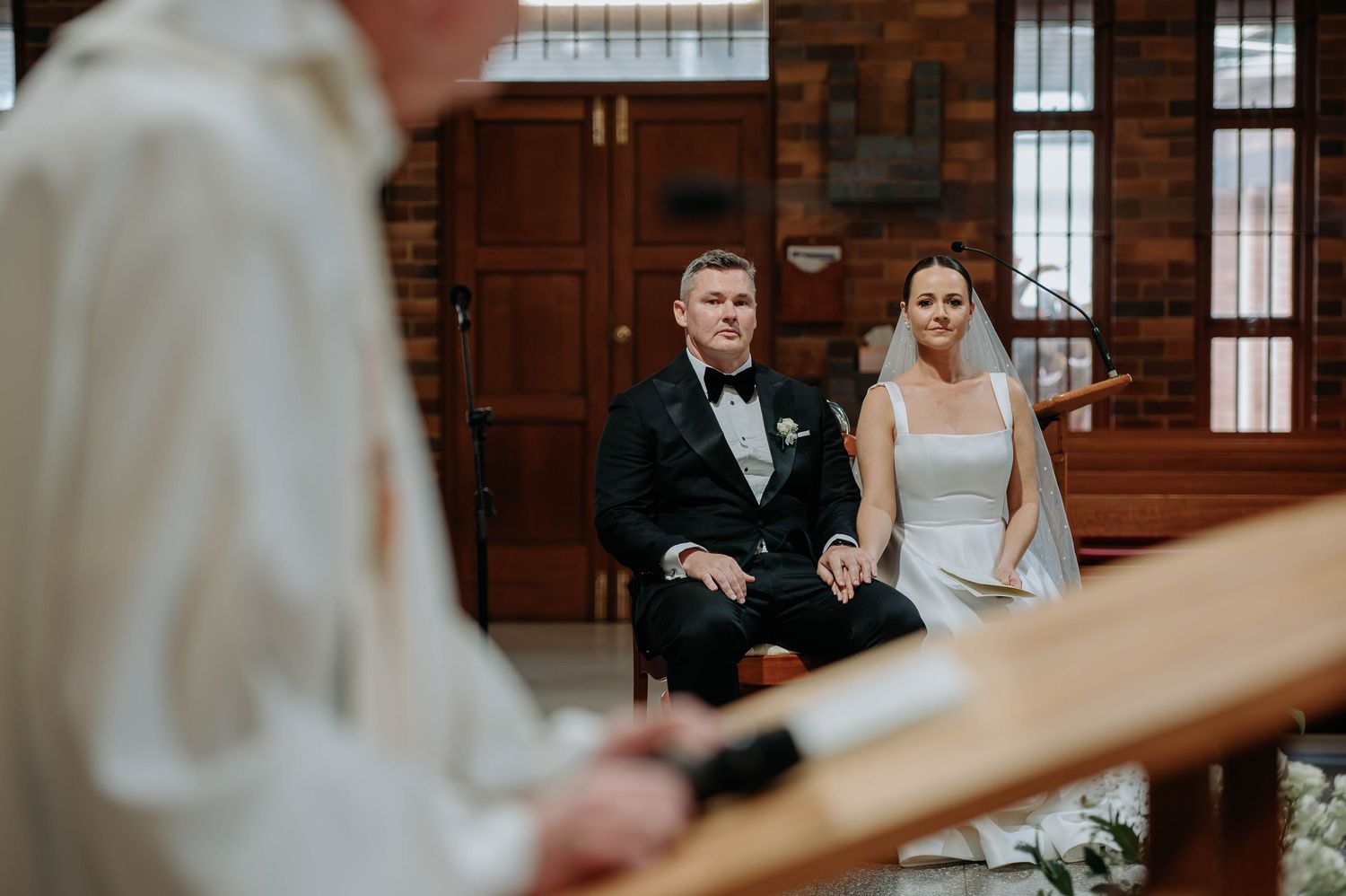 Wedding ceremony in a brick church interior with wooden pews and stained glass windows.