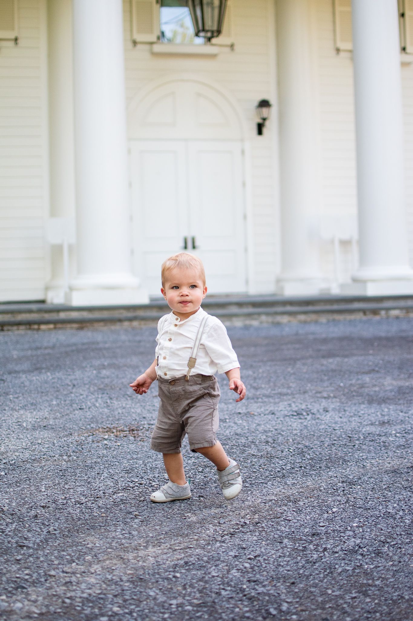 Small child wearing light colored shirt and gray shorts walking on asphalt driveway in front of white pillared building.