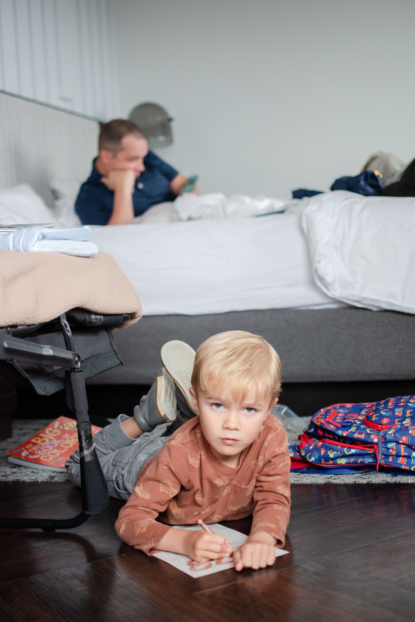 A young child lies on a hardwood floor writing on paper while someone rests on a bed in the background.