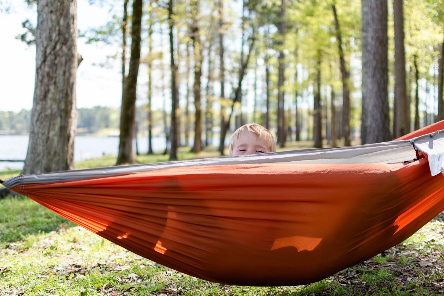 A bright orange camping hammock hangs between trees near a lakeside, creating a peaceful outdoor relaxation spot.