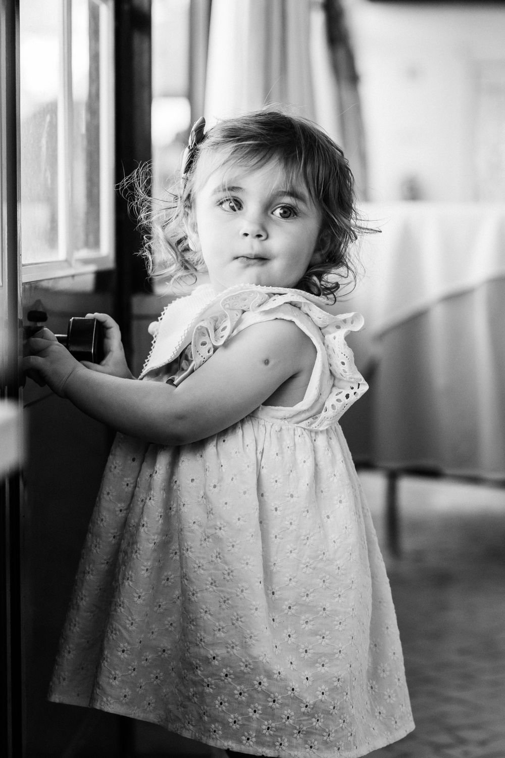 Black and white portrait of a young child in a dotted dress near a window.