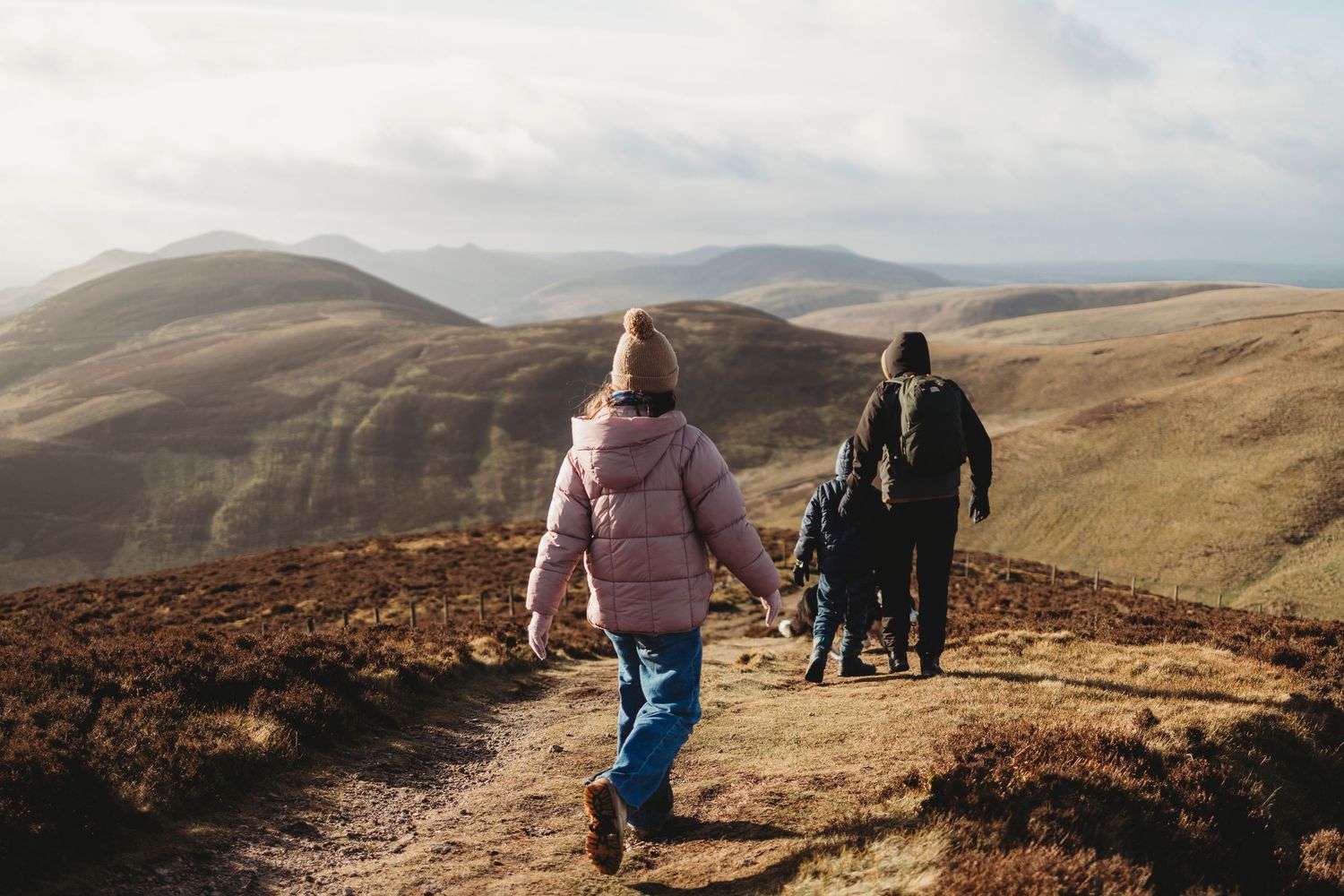 Hikers walking along a scenic mountain trail through rolling brown hills on an overcast day.