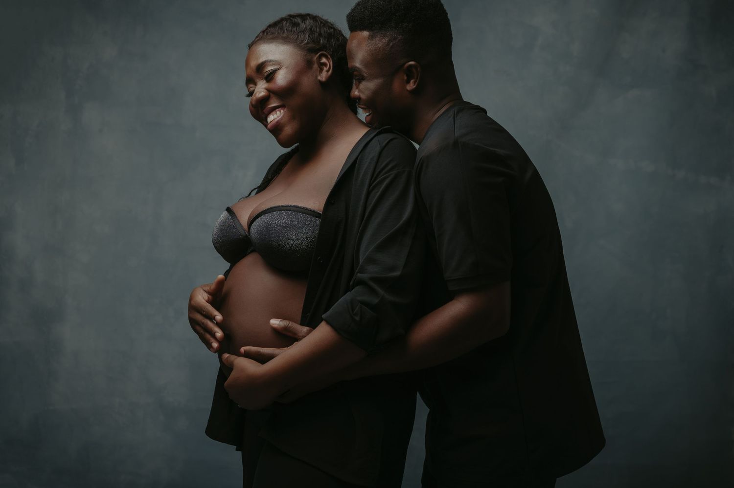 Intimate maternity photoshoot against dark backdrop showing tender moment between couple.