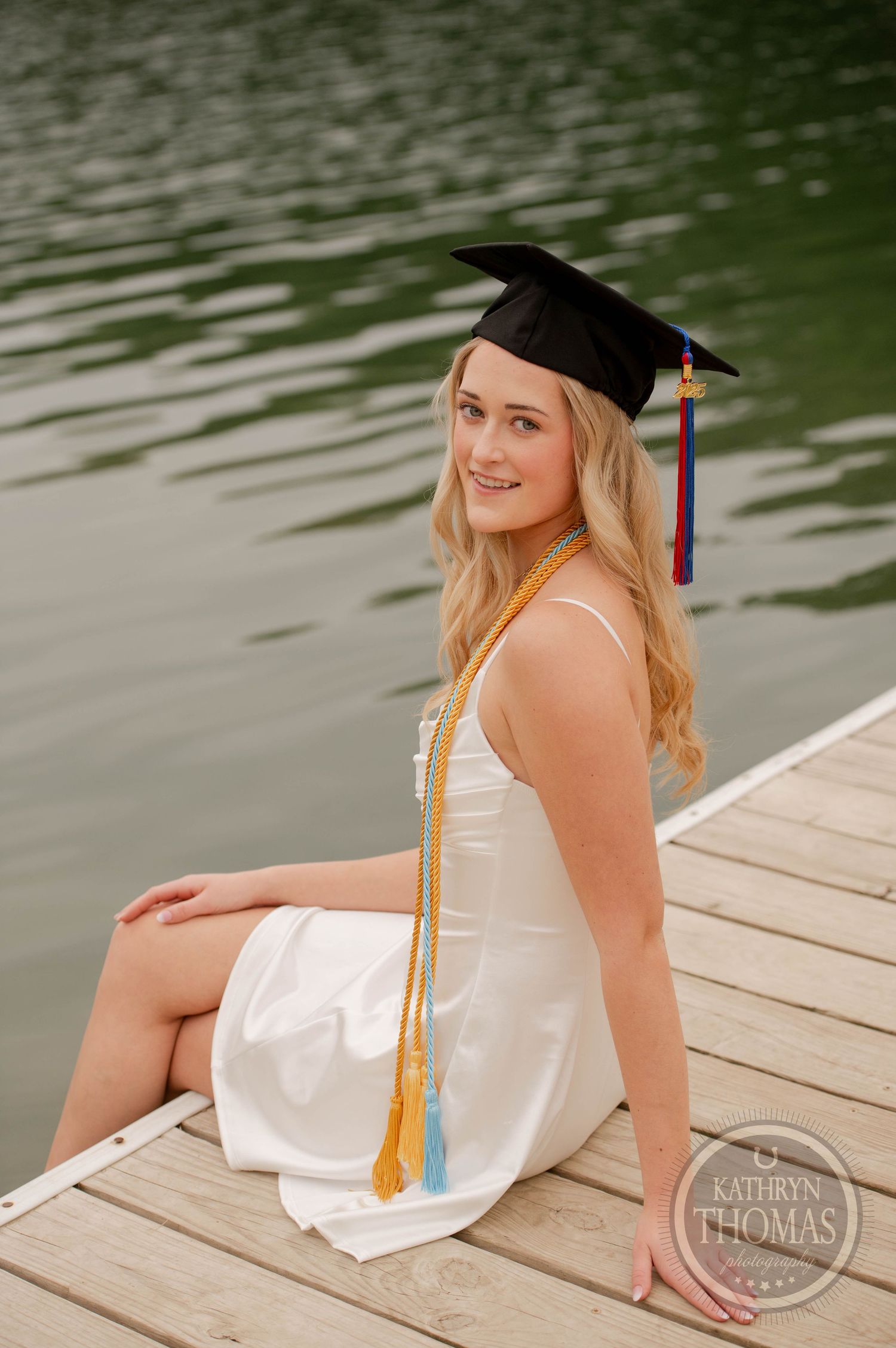 Graduate in white dress and mortarboard cap sits on wooden dock by water with tassel and honor cord.