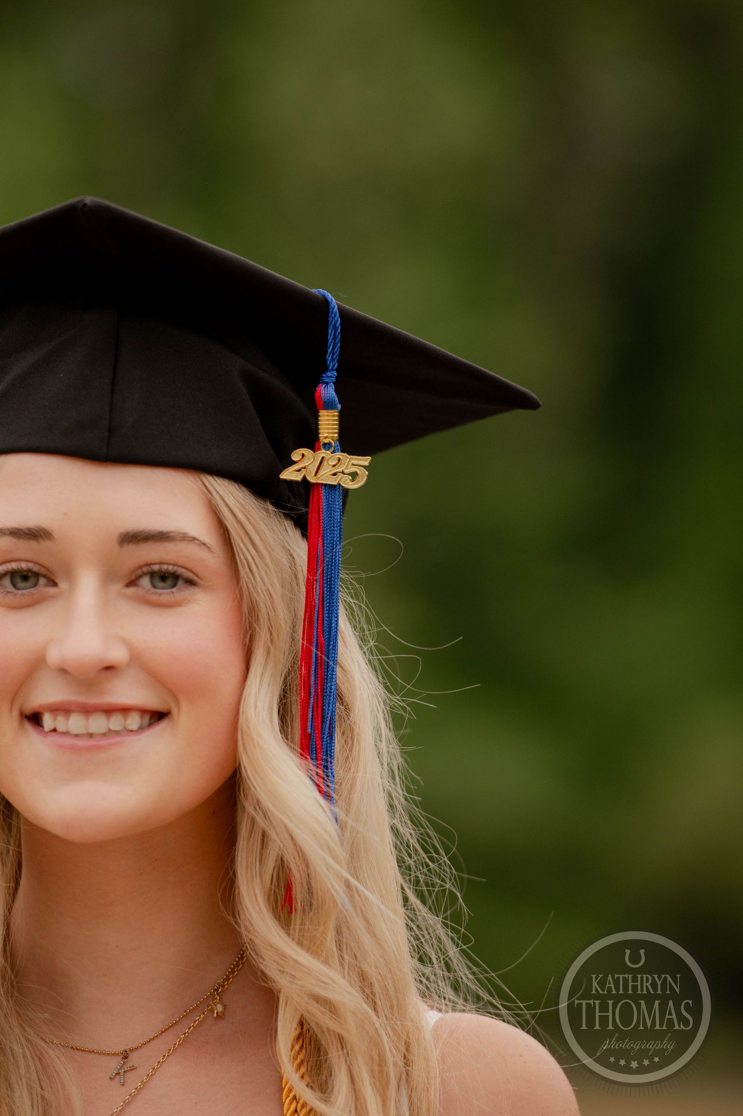 Graduate wearing black mortarboard cap and tassel smiles outdoors against green natural background.