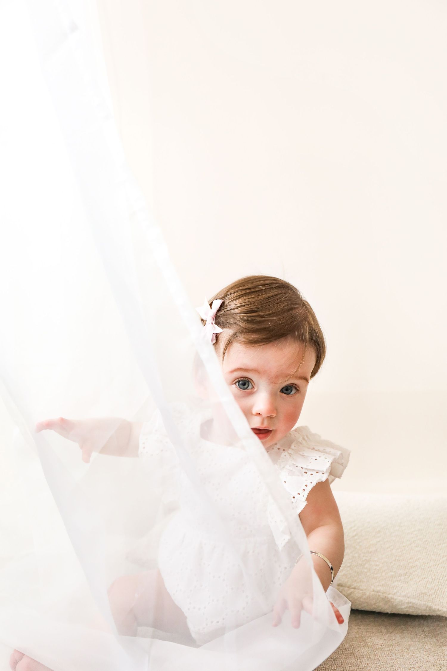 Small child in white lace dress plays with flowing white fabric against light neutral background during Birthday shoot in Nottingham