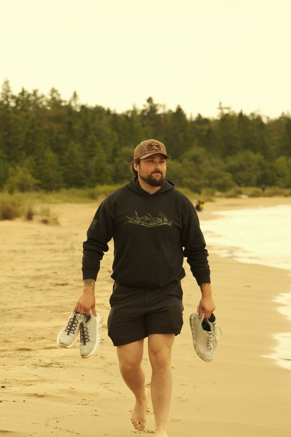 Someone walking barefoot on a beach carrying shoes, wearing black shorts and sweatshirt with a baseball cap at sunset.